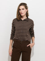 A person stands against a white wall, wearing the Taylor Striped Cardigan in Cotton Cashmere—an oversized brown and beige striped long-sleeve sweater with Troca shell buttons—tucked into high-waisted brown pants, with one hand in a pocket.