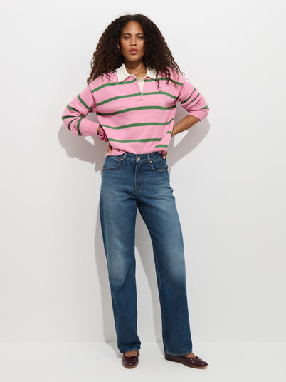A woman with curly hair wears a pink and green striped long-sleeve shirt, the Violet Mid Rise Relaxed Jean in a classic blue wash, and brown shoes, standing against a plain white background with her hands in her pockets.