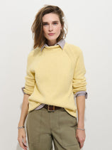 A person with light brown hair wears the Eastport Rollneck In Cotton, a 100% cotton yellow fisherman’s sweater, over a striped shirt and olive green pants, standing against a plain white background and looking at the camera with a neutral expression.